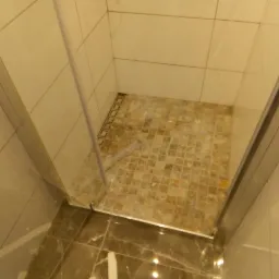 Wnętrze kabiny prysznicowej z mozaikową podłogą i jasnymi płytkami ściennymi, widok z góry, częściowo zamontowane szklane drzwi.