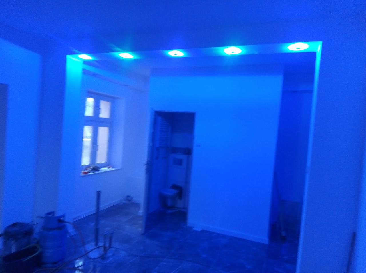 Wnętrze w trakcie remontu, z nową instalacją oświetleniową LED w kolorze niebieskim, widoczne narzędzia i butla gazowa, okno z widokiem na zewnątrz.
