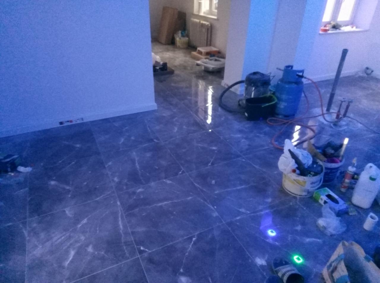 Szare płytki podłogowe imitujące marmur w trakcie montażu, widoczne wiadro z klejem, butla gazowa i odkurzacz przemysłowy, pomieszczenie w trakcie remontu z elementami grzejnika i okna w tle.