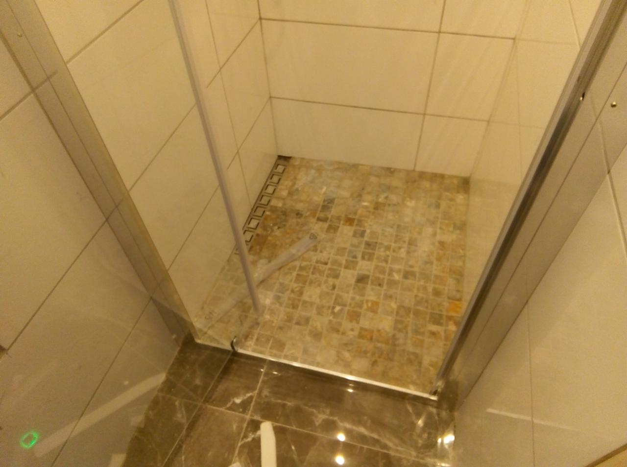 Wnętrze kabiny prysznicowej z mozaikową podłogą i jasnymi płytkami ściennymi, widok z góry, częściowo zamontowane szklane drzwi.