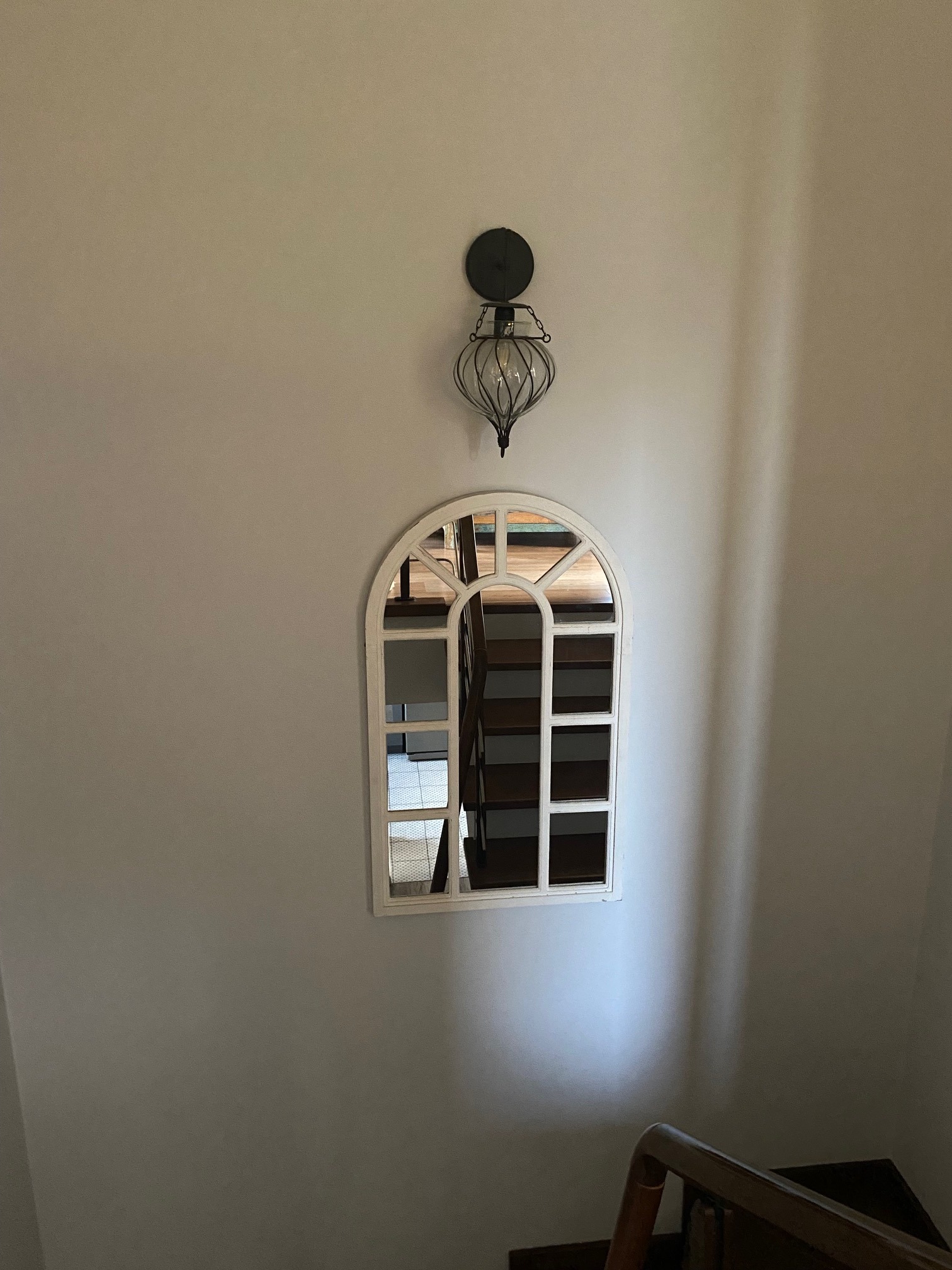 Ściana klatki schodowej z dekoracyjnym lustrem w kształcie okna z białymi szprosami i metalową lampą ścienną w stylu retro