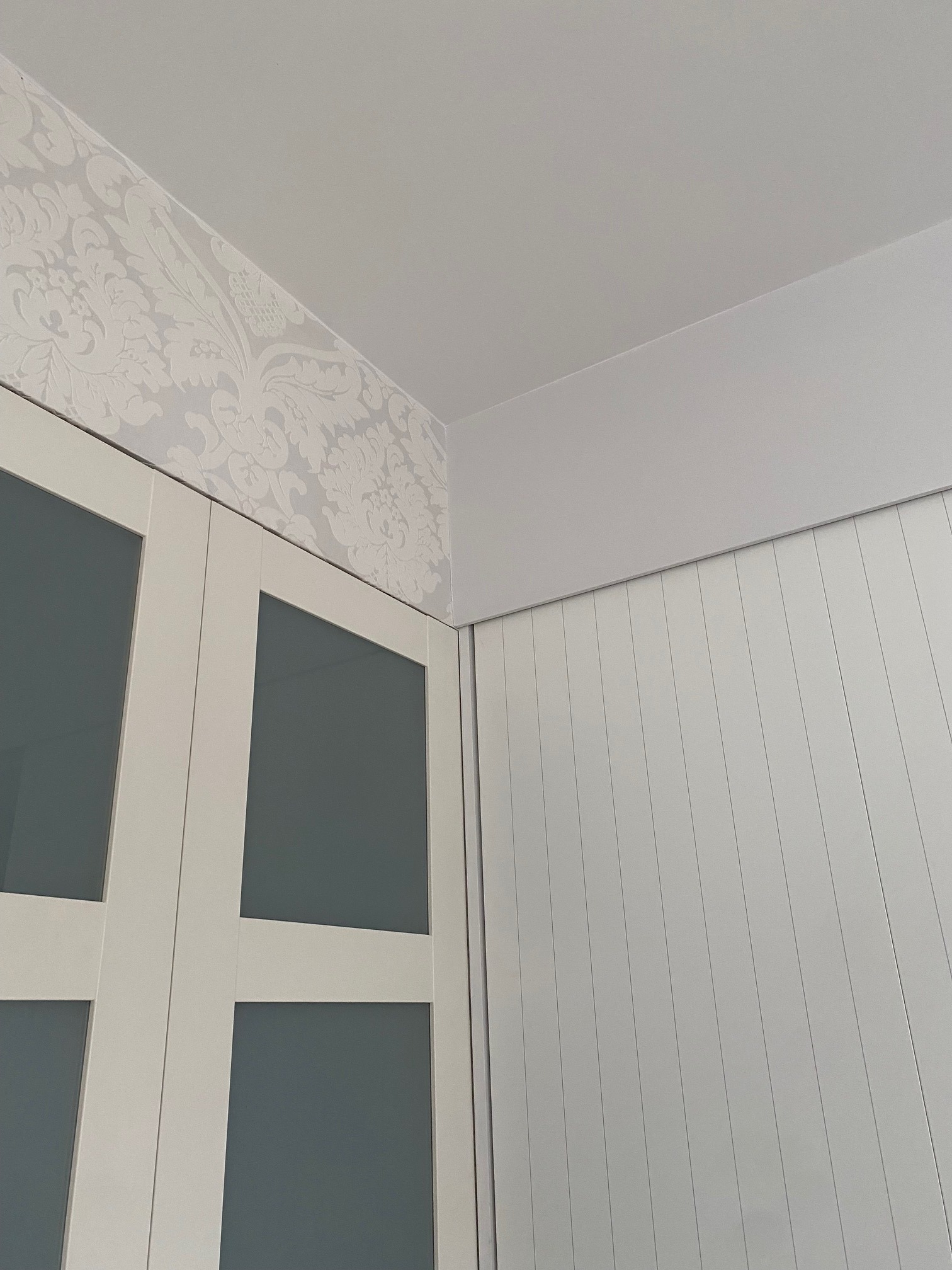 Fragment wnętrza: biała ściana z pionowymi panelami, biała szafa z szarymi szybami i fragment ściany z tapetą w kwiatowy wzór w odcieniach szarości.