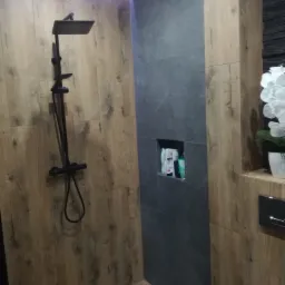 Nowoczesna kabina prysznicowa z deszczownicą w kolorze czarnym, wykończona płytkami imitującymi drewno i szarą ścianą z wnęką na kosmetyki, oświetlenie LED w suficie.