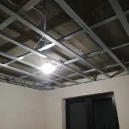 Metalowa konstrukcja sufitu podwieszanego z widocznymi przewodami elektrycznymi przed montażem płyt, oświetlona tymczasową lampą, z fragmentem ściany i czarnych drzwi w tle.