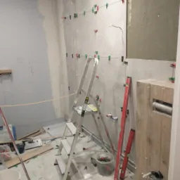 Łazienka w trakcie montażu płytek: widoczne płytki przytwierdzone do ściany za pomocą klipsów poziomujących, drabina aluminiowa, poziomice oraz zabudowa stelaża WC w drewnopodobnej okleinie.