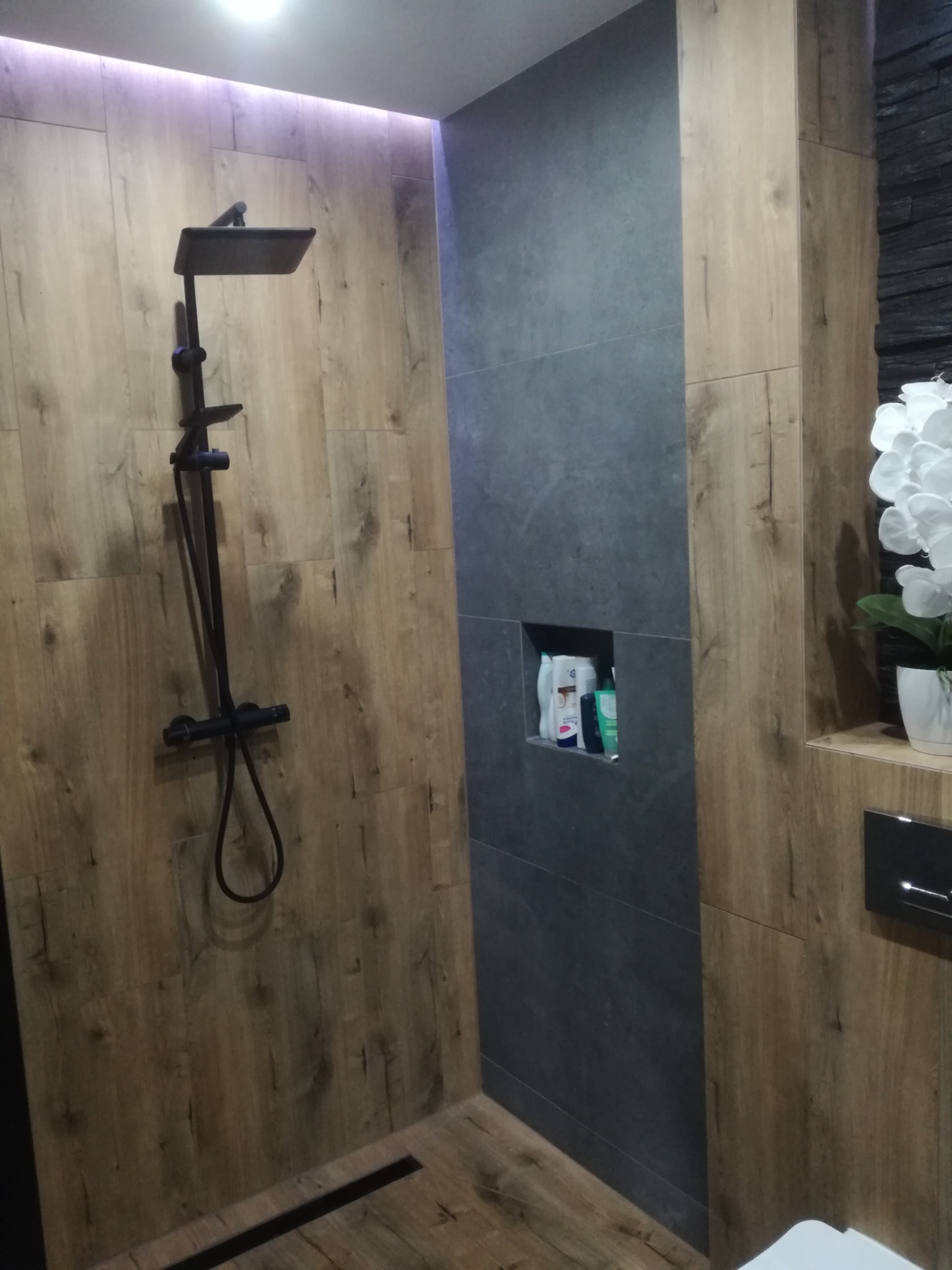 Nowoczesna kabina prysznicowa z deszczownicą w kolorze czarnym, wykończona płytkami imitującymi drewno i szarą ścianą z wnęką na kosmetyki, oświetlenie LED w suficie.