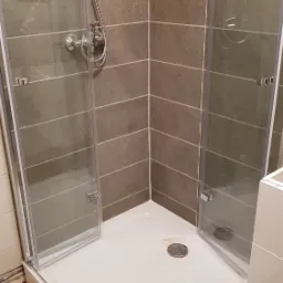 Nowoczesna kabina prysznicowa narożna z szarymi płytkami w poziomym układzie, chromowana bateria i odpływ. Widoczne ślady po montażu na podłodze.