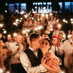 Para młoda w objęciach, otoczona gośćmi weselnymi z zimnymi ogniami w ciemności.