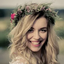 Portret uśmiechniętej kobiety z blond włosami i wiankiem z różowych i białych kwiatów na głowie, delikatny makijaż, widoczny fragment koronkowej sukienki.