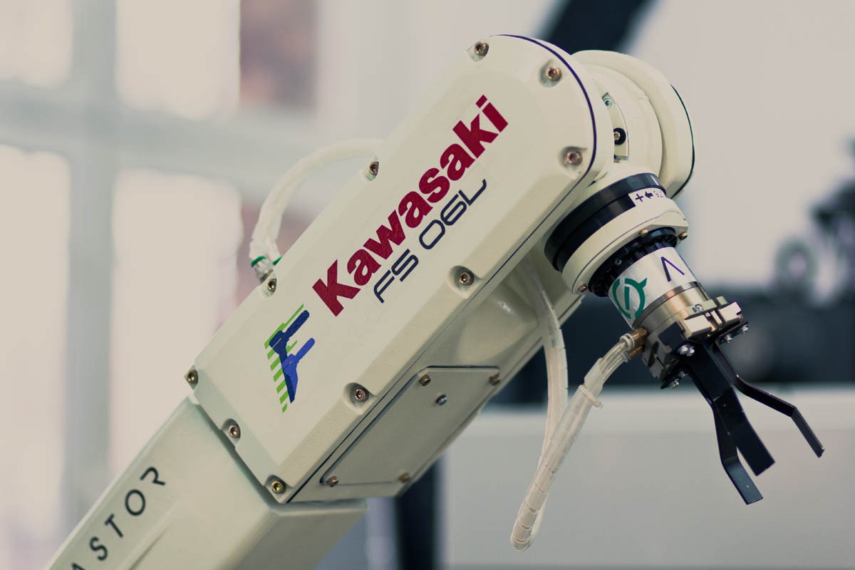 Ramię robota przemysłowego Kawasaki FS 06L z chwytakiem, widoczne przewody i logo producenta na tle okna.