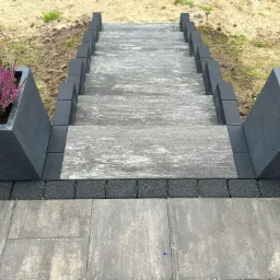 Schody zewnętrzne wyłożone szarą kostką brukową, obramowane ciemnymi obrzeżami, z donicami po bokach obsadzonymi wrzosem.