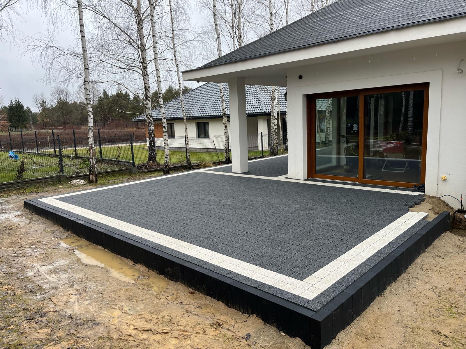 Nowoczesny taras z ciemnoszarej kostki brukowej z białym obramowaniem, przylegający do białego domu z dużymi przesuwnymi drzwiami tarasowymi. Częściowo ułożona nawierzchnia, widoczne niedokończone...