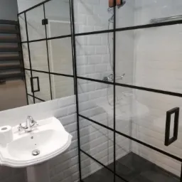 Nowoczesna łazienka z kabiną prysznicową typu walk-in z czarnymi szprosami, białą umywalką na postumencie i grzejnikiem drabinkowym w tle.