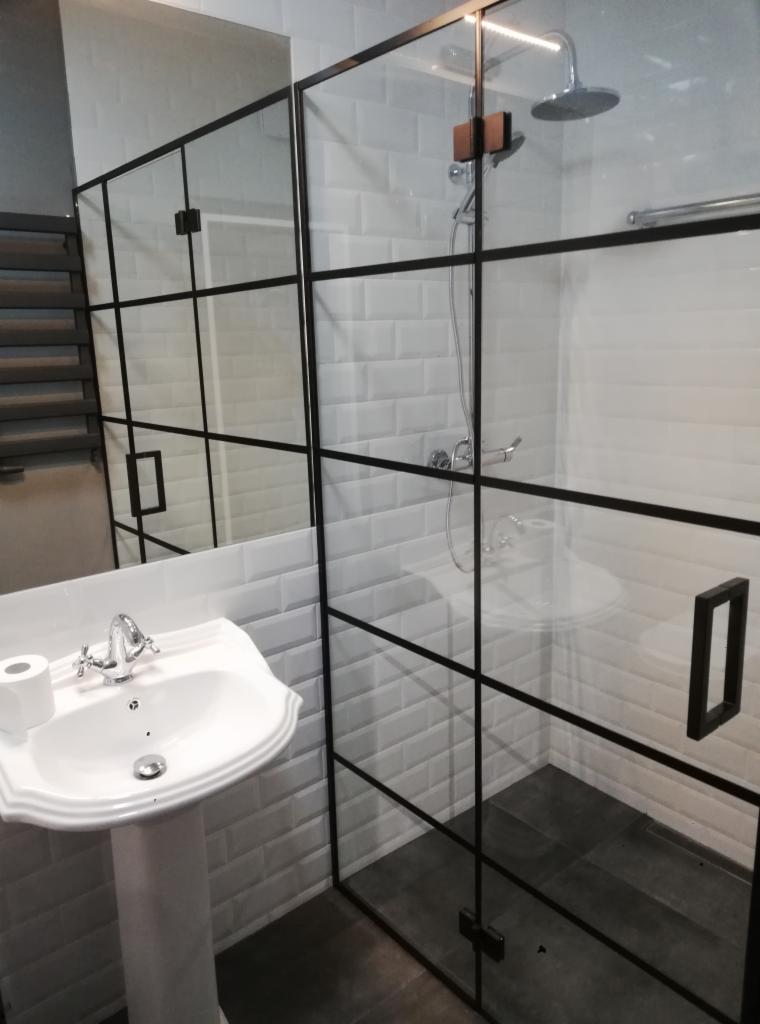 Nowoczesna łazienka z kabiną prysznicową typu walk-in z czarnymi szprosami, białą umywalką na postumencie i grzejnikiem drabinkowym w tle.