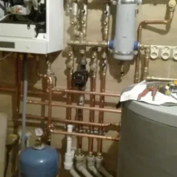 Złożona instalacja centralnego ogrzewania z piecem, zbiornikiem wyrównawczym, pompą obiegową i rurami miedzianymi, widoczne narzędzia na blacie.