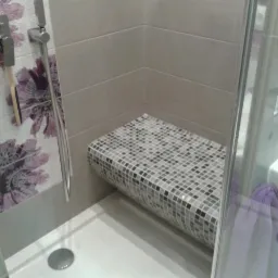 Nowoczesna kabina prysznicowa z siedziskiem wyłożonym mozaiką w odcieniach szarości, ściany wykończone płytkami w kolorach beżu i bieli z motywem kwiatowym w odcieniach fioletu, widoczna bateria...