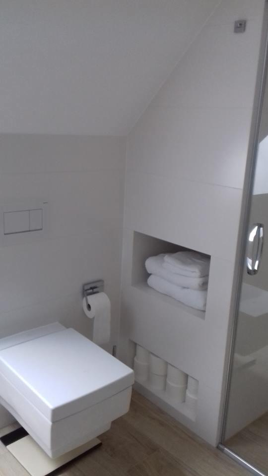 Nowoczesna, minimalistyczna łazienka w odcieniach bieli z podwieszaną toaletą, ukrytym spłukiwaniem, półką na ręczniki i zapas papieru toaletowego w zabudowie oraz kabiną prysznicową ze szklanymi...