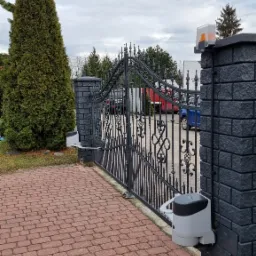 Otwarta, kuta brama wjazdowa z automatyką; widoczny napęd bramy zamontowany na ceglanym słupku, z sygnalizatorem ostrzegawczym na drugim słupku, czerwona kostka brukowa na podjeździe, w tle...