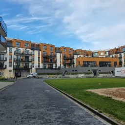 Nowoczesny budynek apartamentowy z drewnianymi akcentami i szklanymi balkonami, widok z poziomu brukowanej drogi w kierunku trawnika i placu budowy.