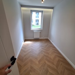 Usługi Budowlane BRT-MOLUS Daniel Molus - Pusty pokój po remoncie z parkietem ułożonym w jodełkę, grzejnikiem pod oknem i widokiem na budynek z balkonami.