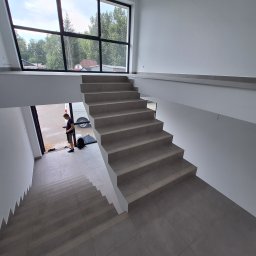 Usługi Budowlane BRT-MOLUS Daniel Molus - Wykończone schody wewnętrzne z szarych płytek z antypoślizgowymi paskami; widoczny fragment okna z widokiem na zewnątrz i mężczyzna w drzwiach.