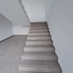 Schody wewnętrzne wyłożone szarymi płytkami ceramicznymi z ryflowanymi krawędziami, dopasowane do szarej podłogi i białych ścian w nowym budynku.