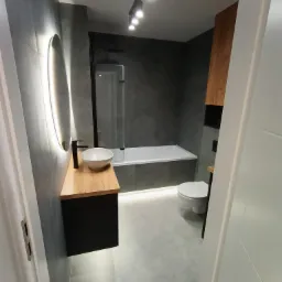 Nowoczesna łazienka w odcieniach szarości z drewnianym blatem pod umywalką, okrągłe lustro z podświetleniem LED, biała wanna z parawanem, podświetlenie podłogowe i czarna armatura.