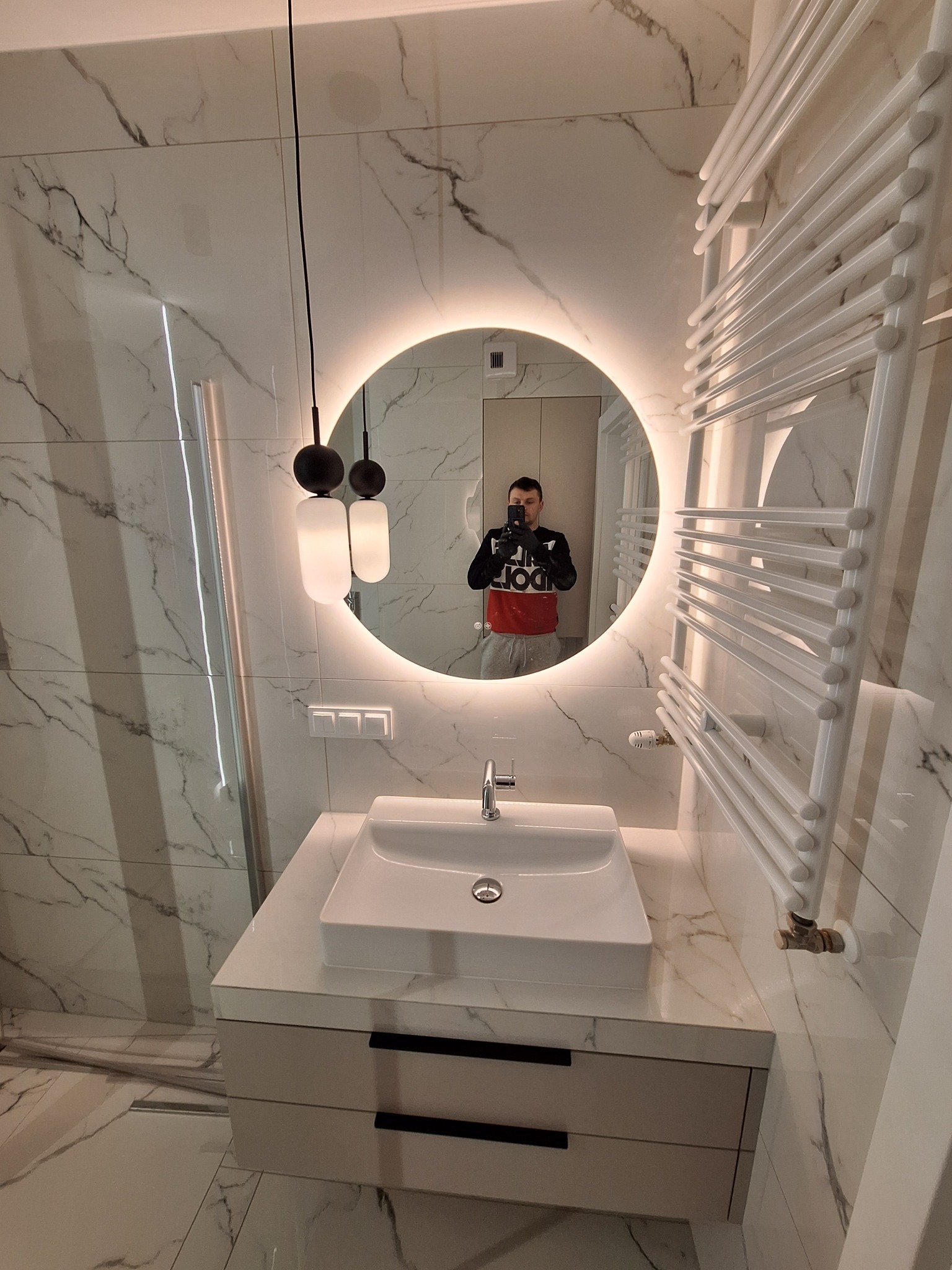 Nowoczesna łazienka z umywalką nablatową na szafce z szufladami, okrągłe lustro z podświetleniem LED, grzejnik drabinkowy i marmurowe płytki na ścianach, widoczne odbicie osoby robiącej zdjęcie.