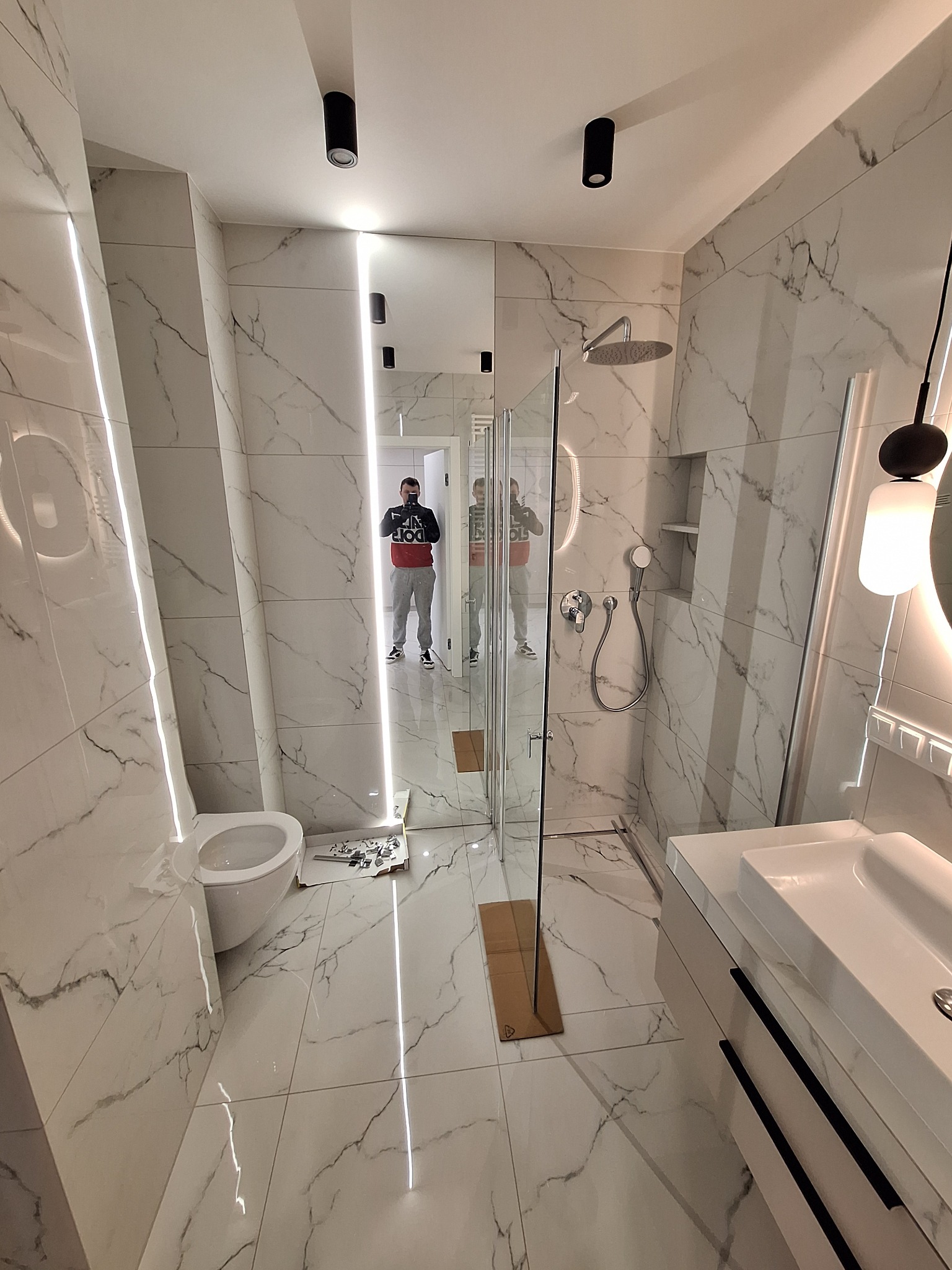 Nowoczesna łazienka wykończona białymi płytkami imitującymi marmur, z kabiną prysznicową typu walk-in, podwieszaną toaletą i umywalką z szafką, oświetlona ledowymi listwami i czarnymi reflektorami...