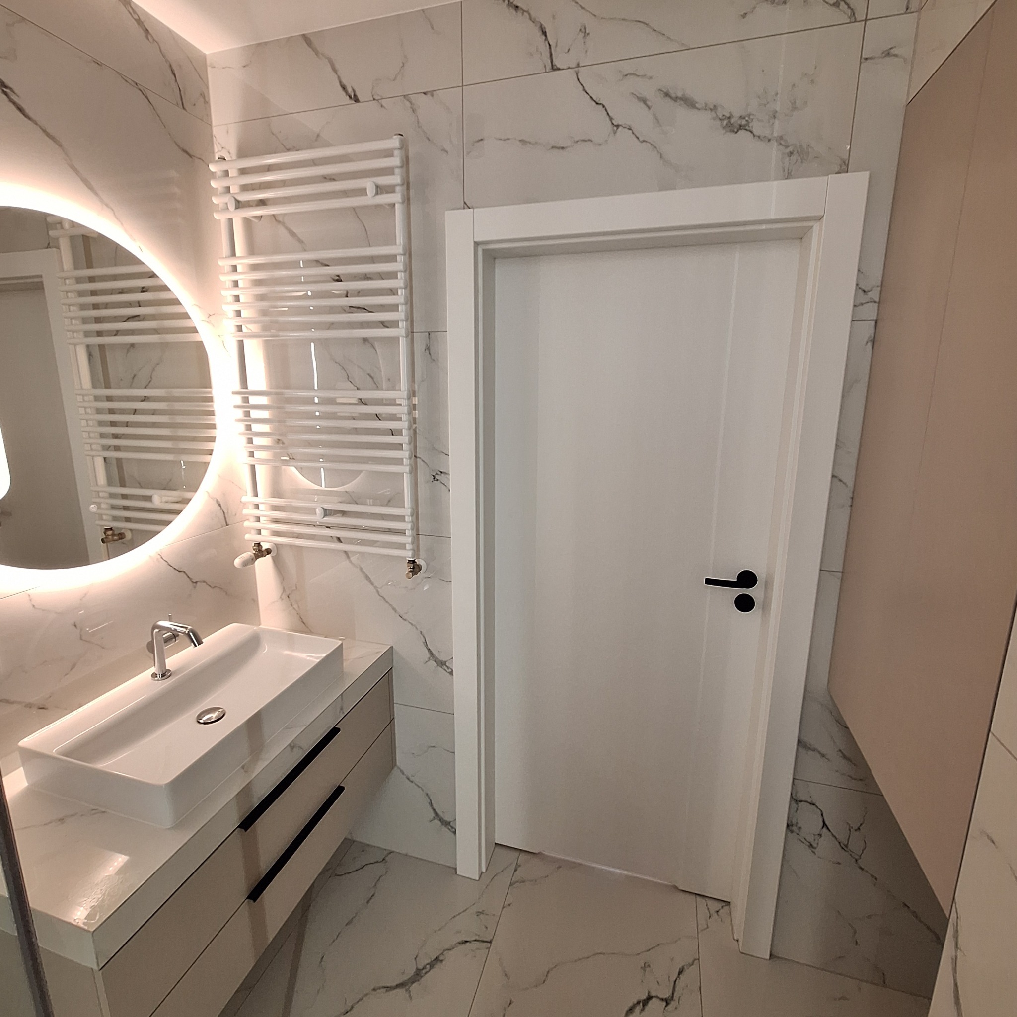 Nowoczesna łazienka z białymi płytkami imitującymi marmur, podświetlanym okrągłym lustrem, białą umywalką nablatową z czarnymi uchwytami, białym grzejnikiem drabinkowym i białą, gładką ścianą.