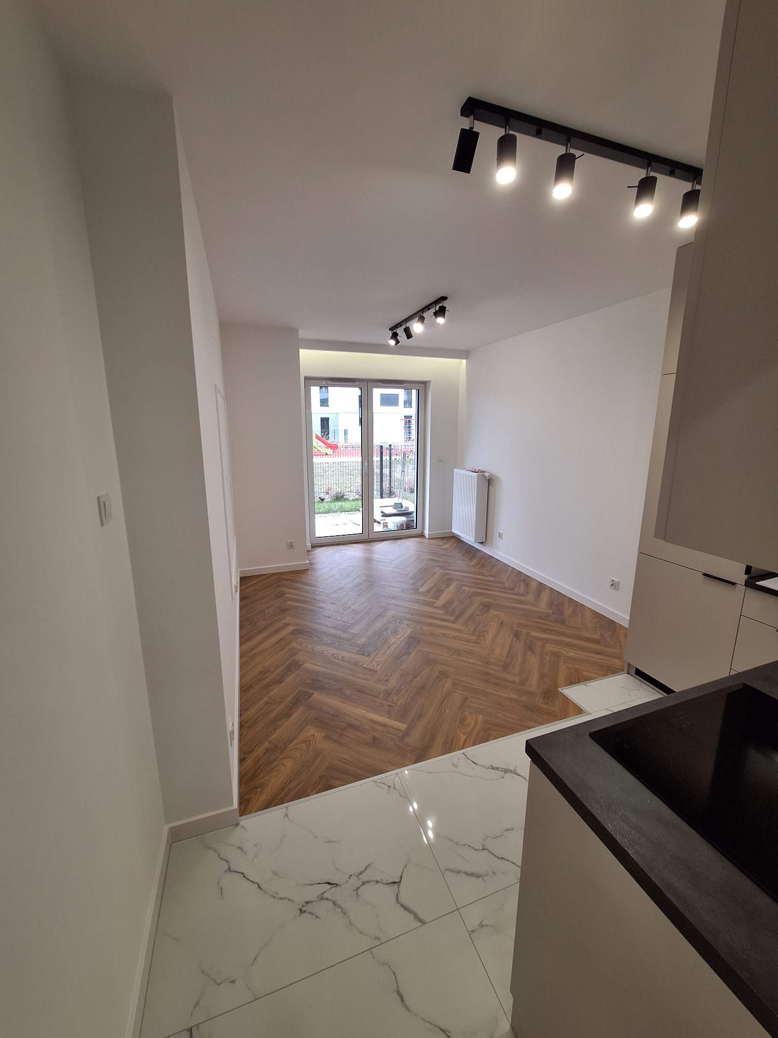 Nowoczesne wnętrze mieszkania po remoncie: parkiet ułożony w jodełkę, białe ściany, oświetlenie szynowe i marmurowe płytki w kuchni.