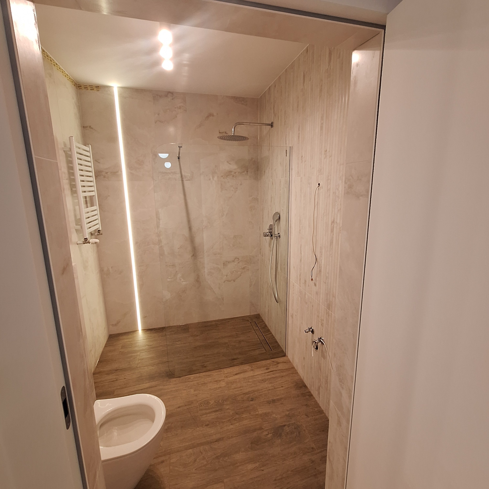 Nowoczesna łazienka z kabiną prysznicową typu walk-in, wykończona płytkami imitującymi drewno i kamień, oświetleniem LED, grzejnikiem drabinkowym i toaletą.