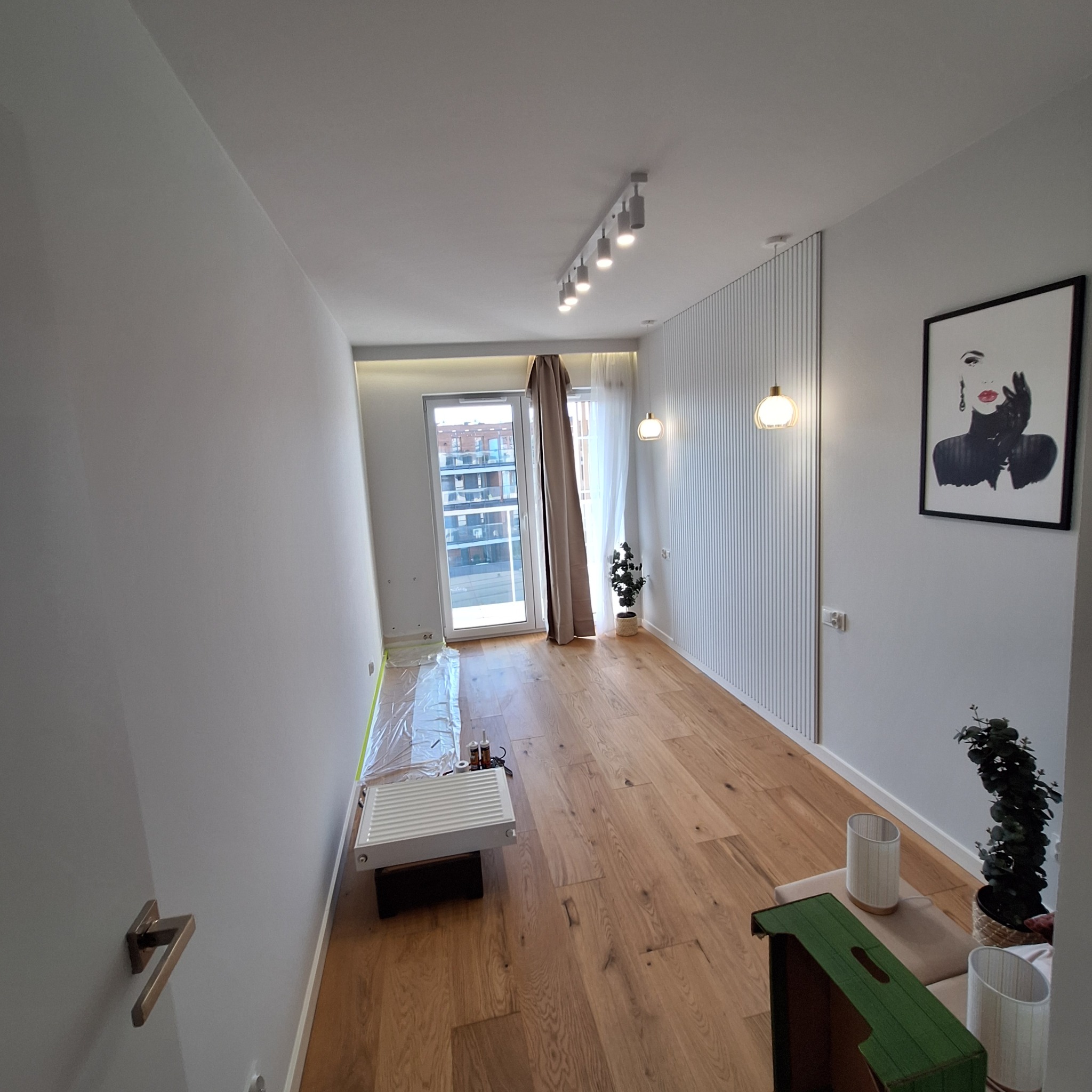 Salon po remoncie w Tokarni z drewnianą podłogą, białymi ścianami z dekoracyjnymi lamelami, nowoczesnym oświetleniem i osłoniętym grzejnikiem.