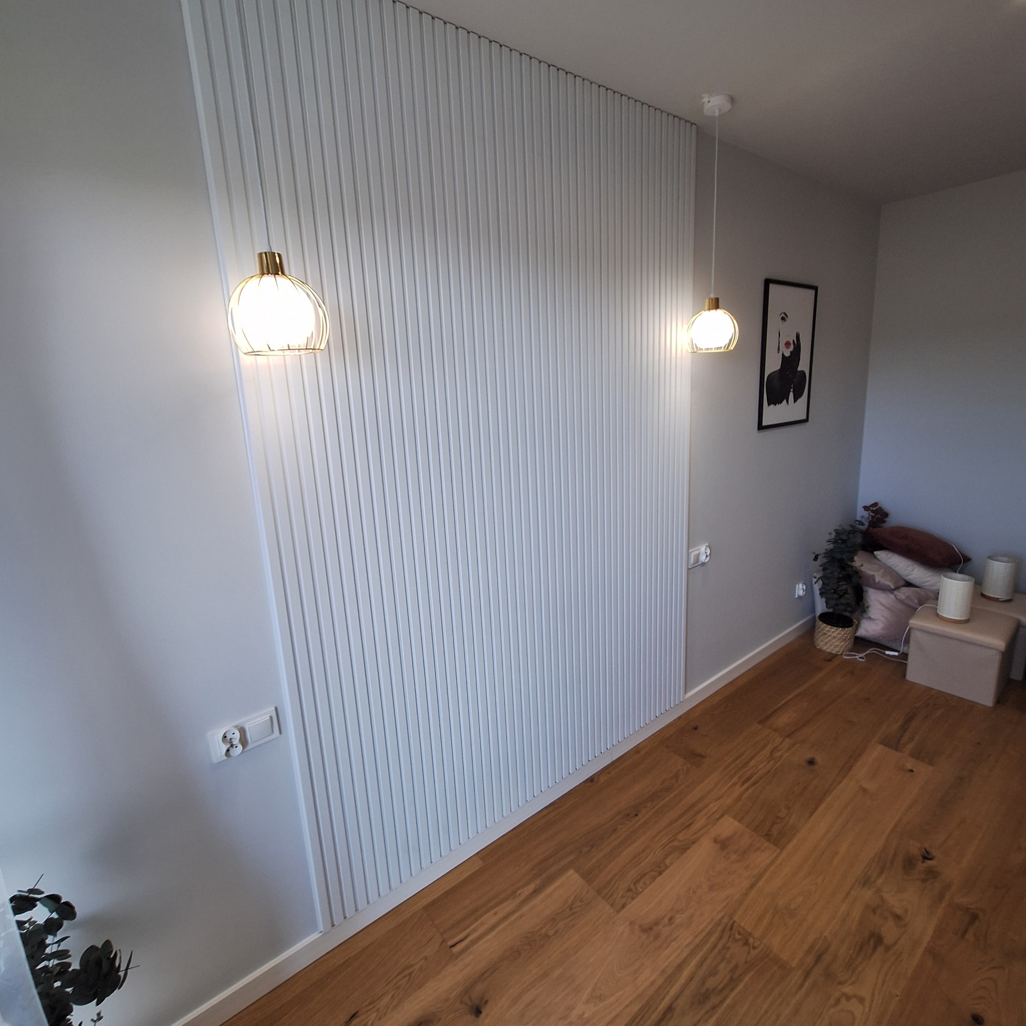 Ściana z pionowych paneli dekoracyjnych w pokoju z drewnianą podłogą, oświetlona dwoma wiszącymi lampami z ażurowymi kloszami, z gniazdkami elektrycznymi na ścianie i minimalistycznym obrazem w tle.
