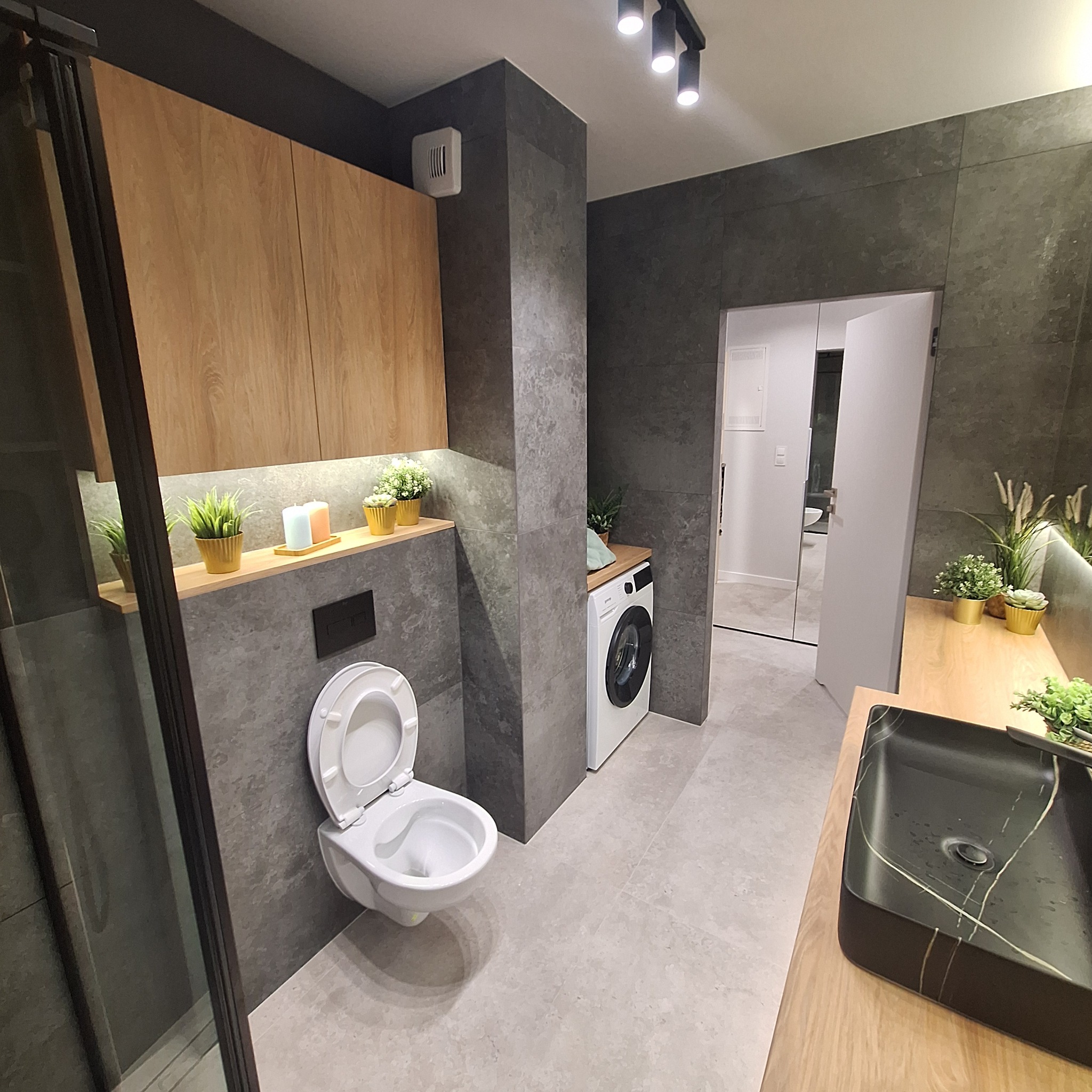 Nowoczesna łazienka z szarymi płytkami, podwieszaną toaletą z otwartą klapą, pralką, czarną umywalką nablatową z marmurowym wzorem i drewnianym blatem, oświetleniem punktowym oraz lustrem w tle...