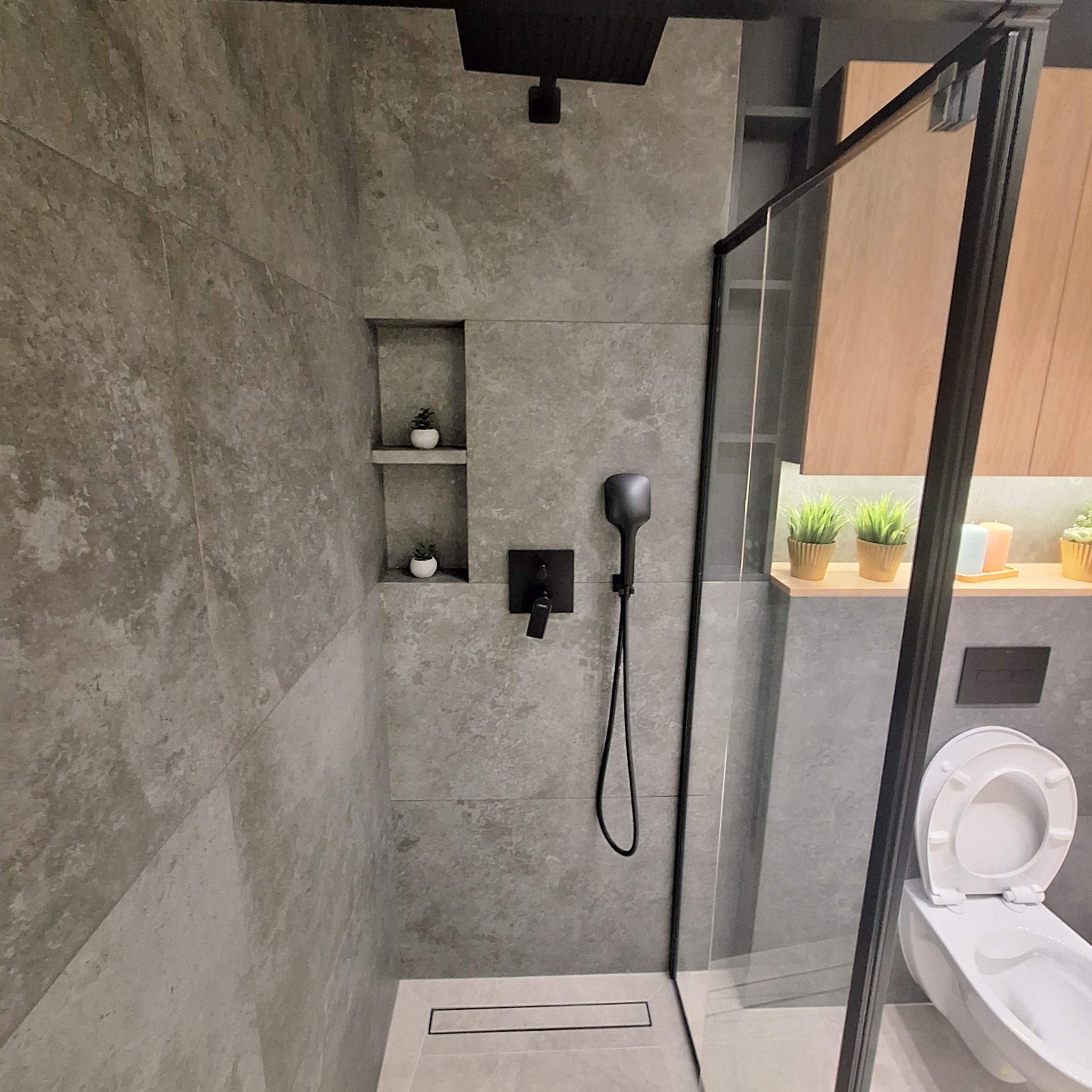 Nowoczesna kabina prysznicowa z szarymi płytkami imitującymi kamień, czarną armaturą, odpływem liniowym w podłodze oraz wnękami z dekoracyjnymi roślinami. Za szklaną ścianką widoczna toaleta...
