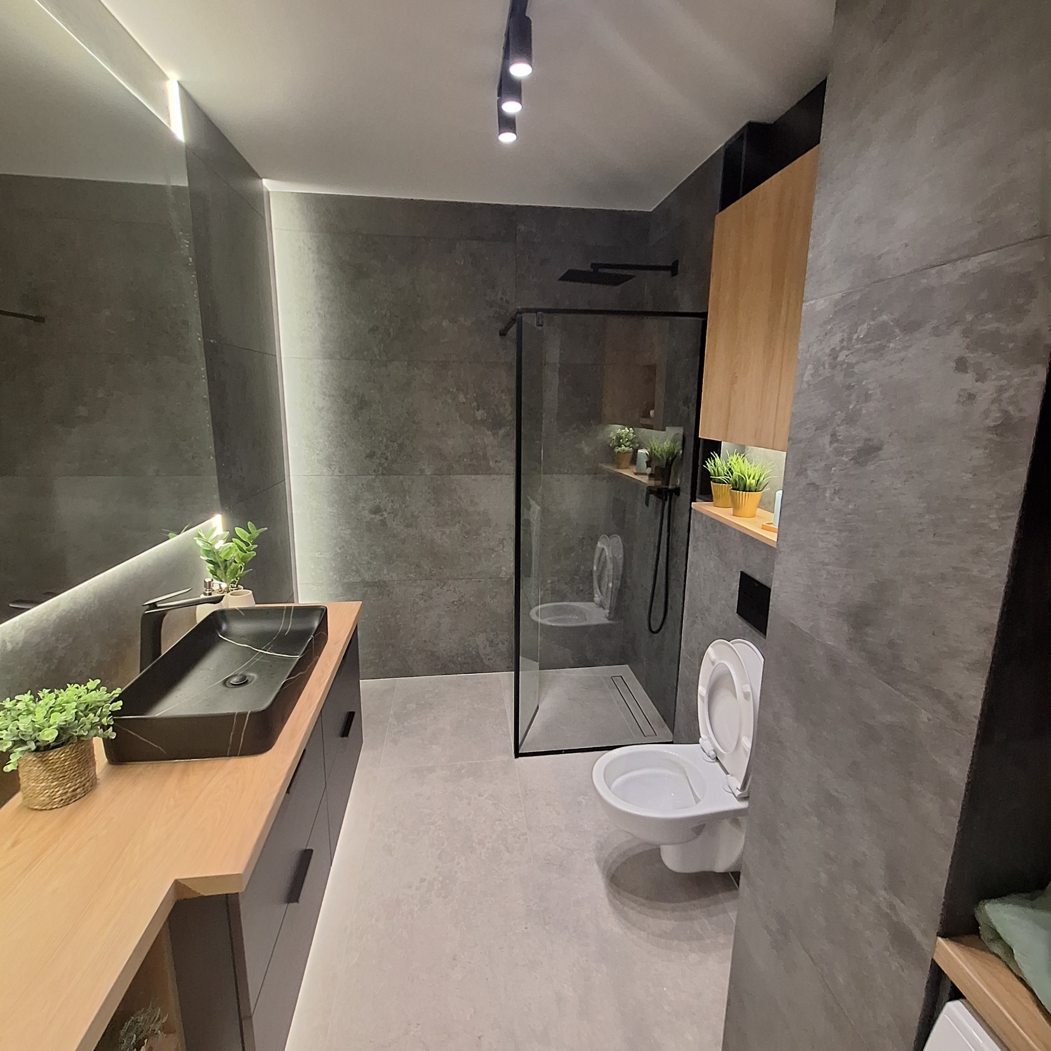Nowoczesna łazienka z szarymi płytkami, czarną umywalką na drewnianym blacie, kabiną prysznicową typu walk-in i podświetleniem LED w lustrze. Uwagę zwraca wisząca szafka z drewnianym frontem...