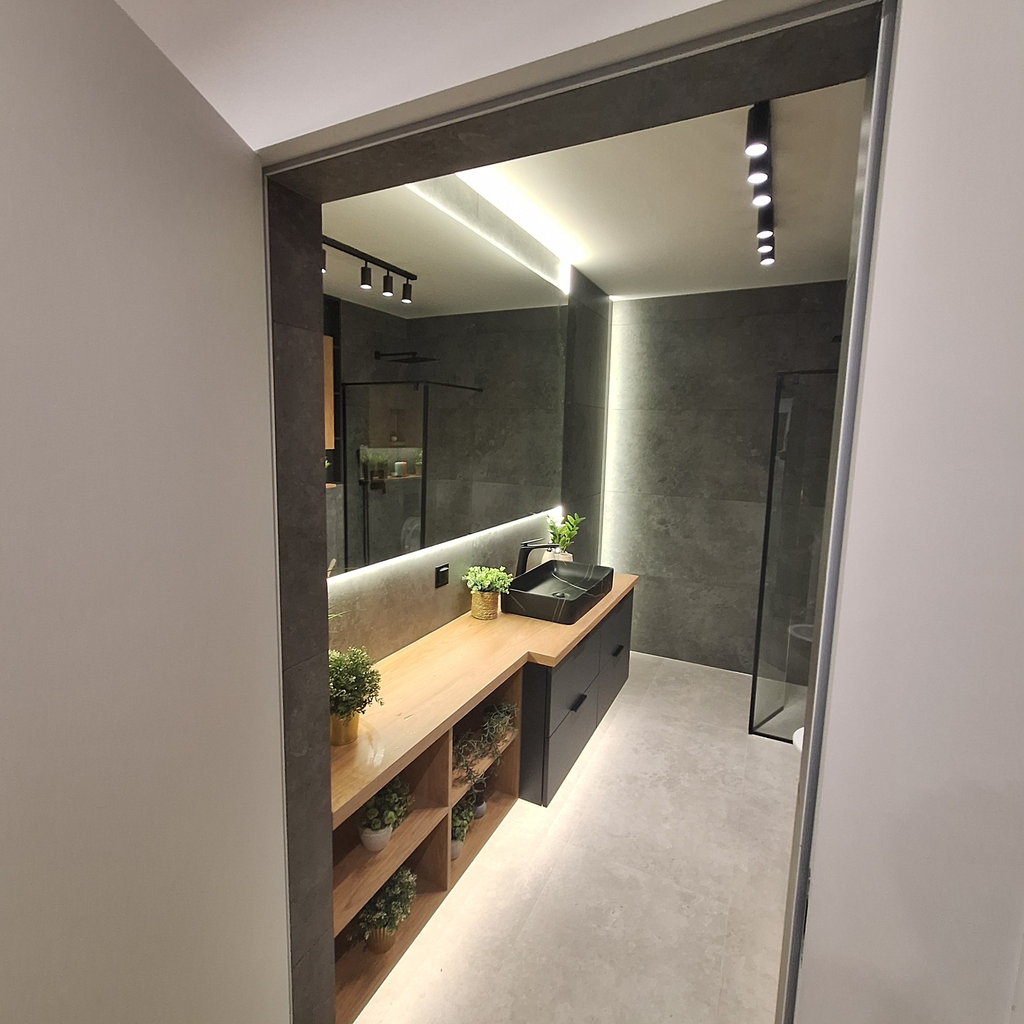 Nowoczesna łazienka z szarą mozaiką na ścianach, podświetlanym lustrem nad drewnianym blatem z czarną umywalką i szafkami, dodatkowo półki z dekoracyjnymi roślinami doniczkowymi.
