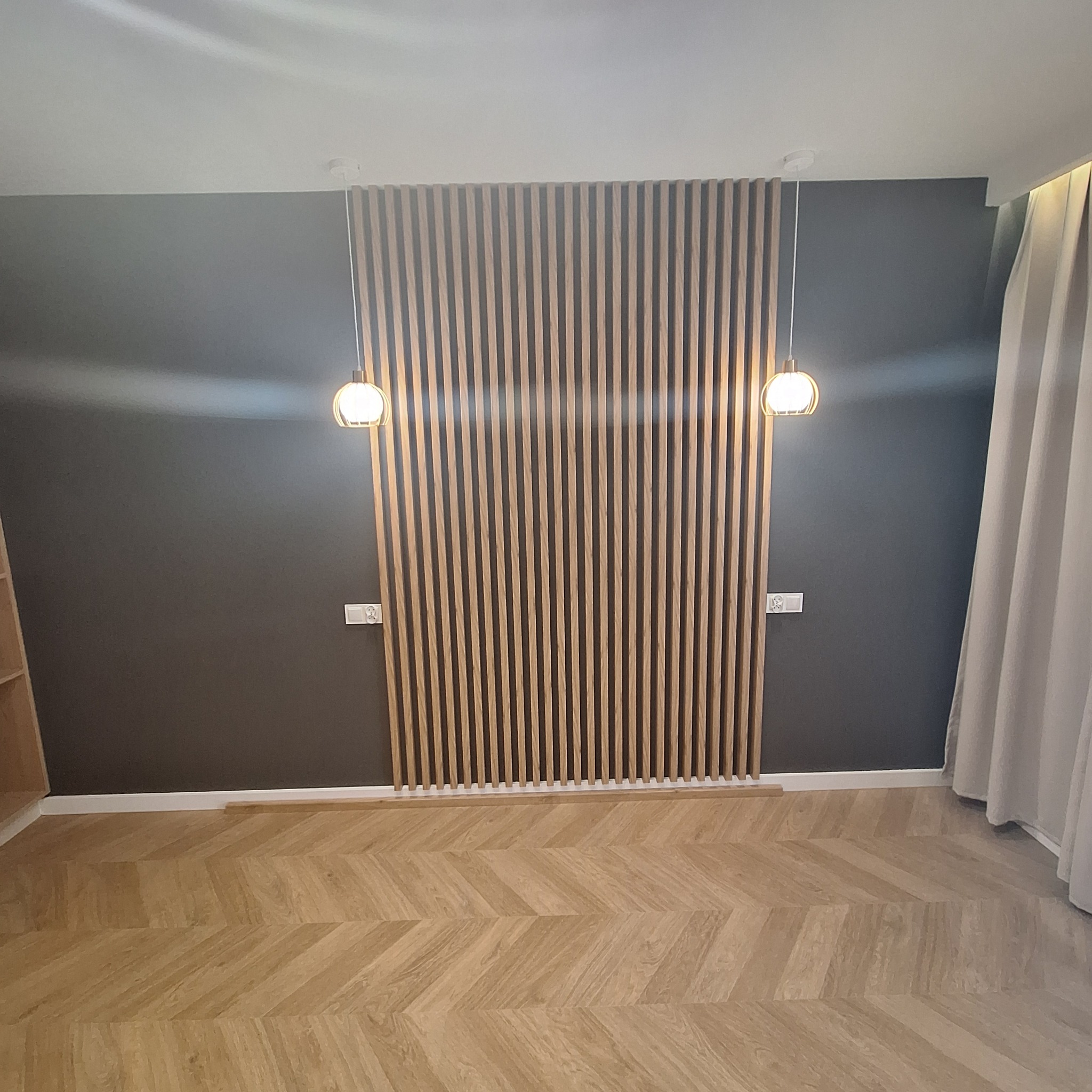 Pokój z drewnianą ścianką działową z pionowych listew, parkietem ułożonym w jodełkę, szarą ścianą i wiszącymi kulistymi lampami po bokach ścianki.