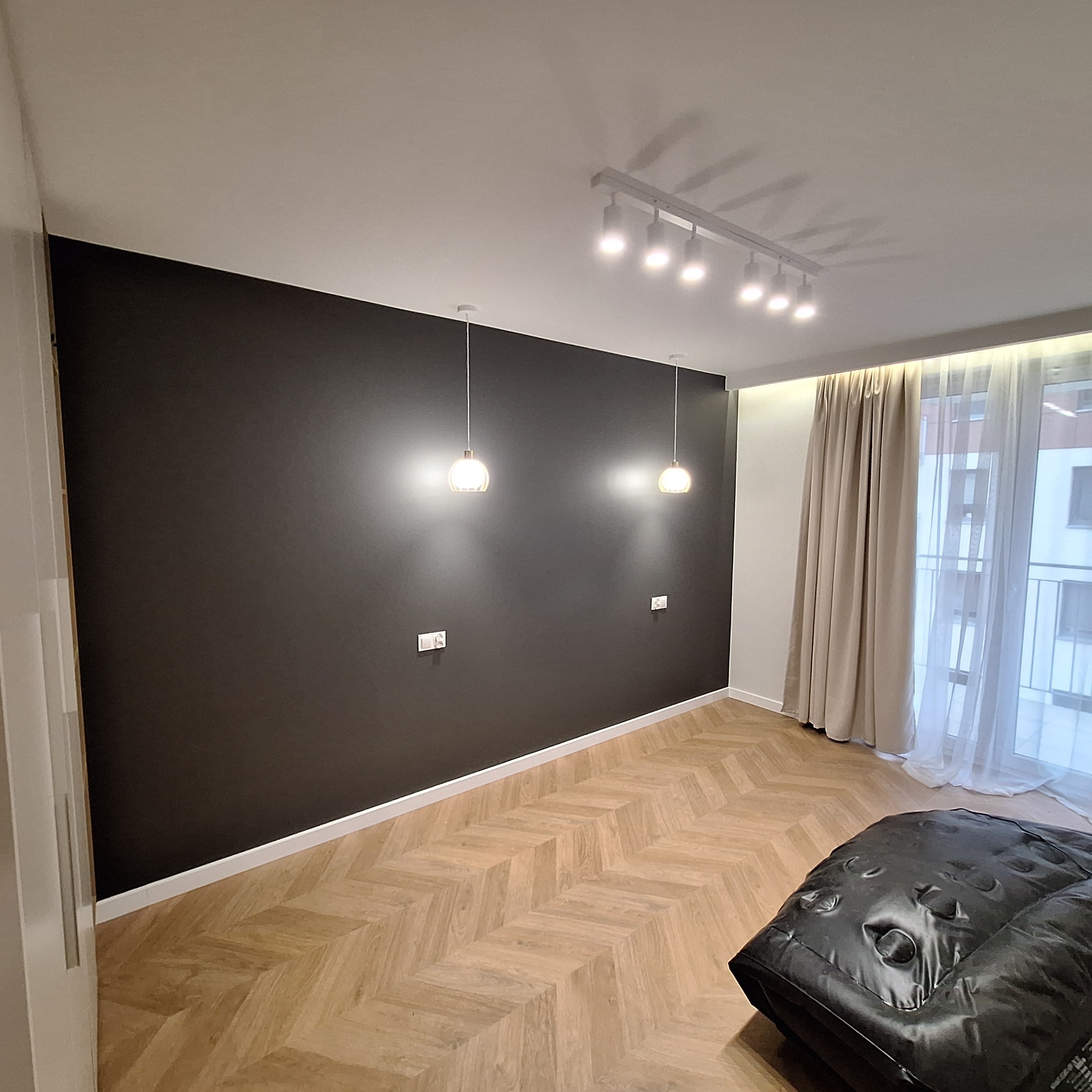 Nowoczesne wnętrze z ciemną ścianą, parkietem w jodełkę, wiszącymi lampami i listwą sufitową z oświetleniem punktowym, widok na okno balkonowe z beżowymi zasłonami.