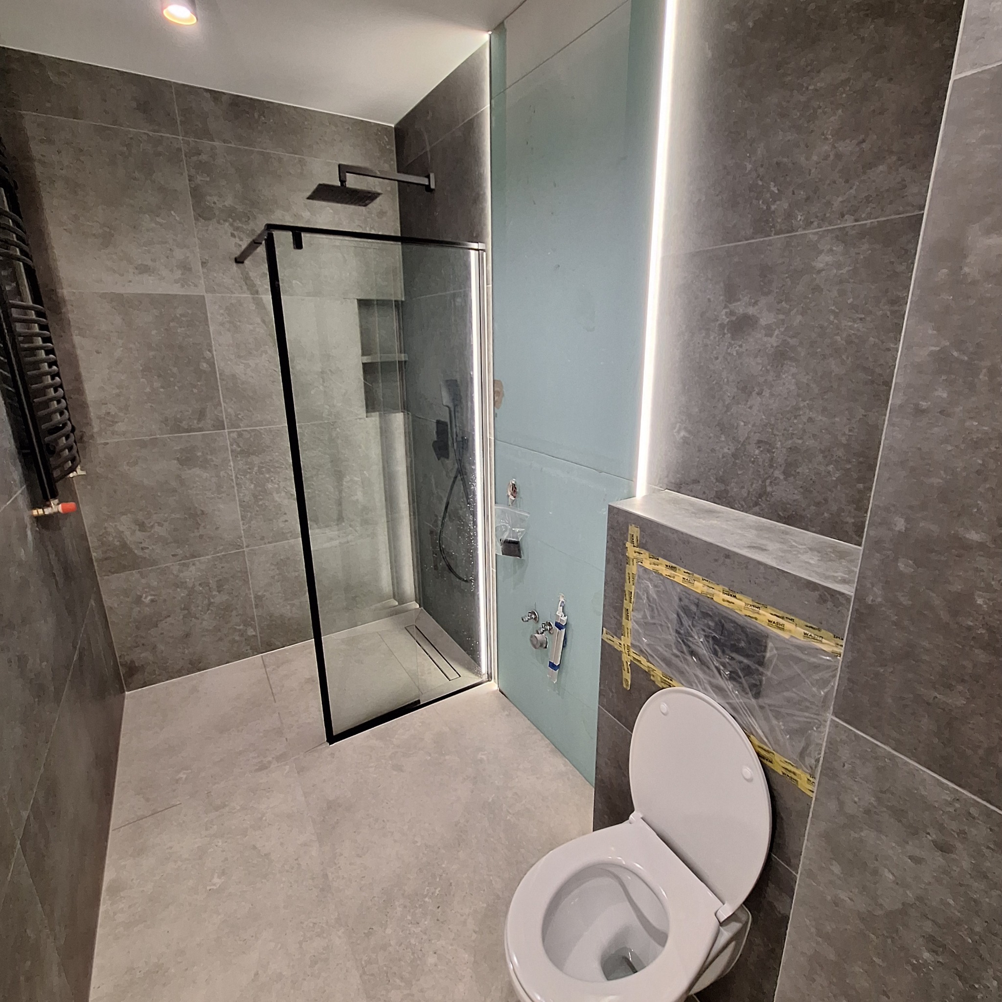 Nowoczesna łazienka z szarymi płytkami, kabiną prysznicową typu walk-in z czarnymi profilami, wiszącą toaletą i grzejnikiem drabinkowym. Fragment ściany z instalacją wodną zabezpieczony folią i taśmą.
