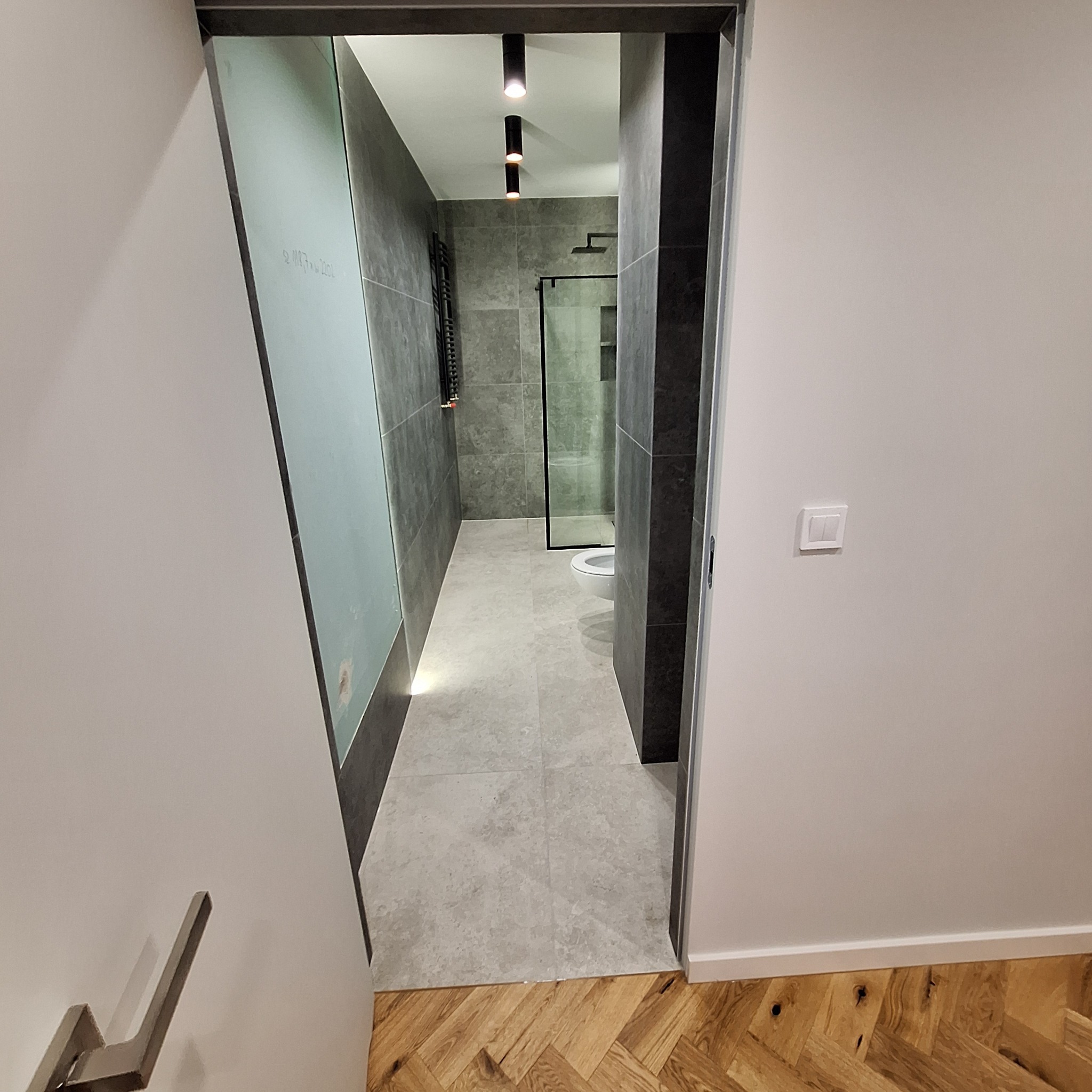 Wnętrze łazienki w trakcie remontu, widoczne szare płytki na ścianach i podłodze, czarna kabina prysznicowa, grzejnik drabinkowy, sufitowe lampy punktowe i częściowo położona podłoga z parkietu...