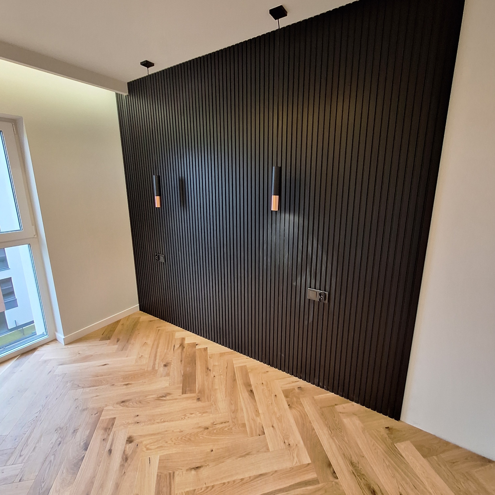 Nowoczesne wnętrze z drewnianą podłogą w jodełkę i ścianą z pionowych, czarnych paneli dekoracyjnych z wbudowanymi lampami i gniazdkami elektrycznymi.