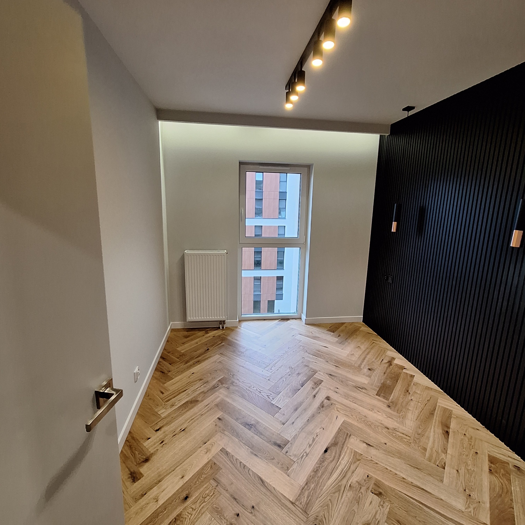 Jasny parkiet ułożony we wzór jodełki francuskiej, szara ściana, czarna ściana z pionowych paneli, okno z widokiem na budynek, nowoczesne oświetlenie szynowe.