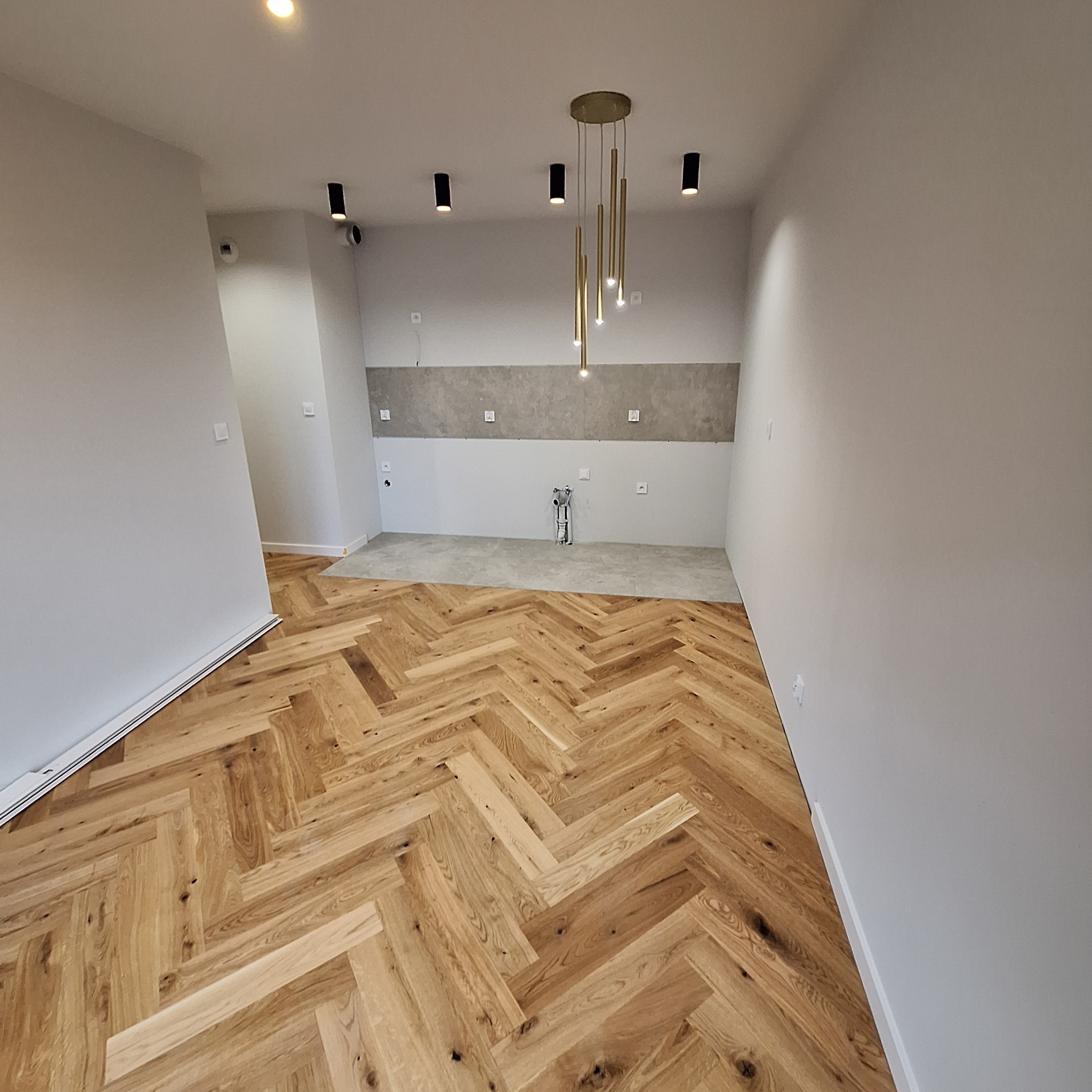 Parkiet w jodełkę francuską z jasnego drewna w nowym wnętrzu, widoczne przygotowane ściany pod zabudowę kuchenną i minimalistyczne oświetlenie punktowe.