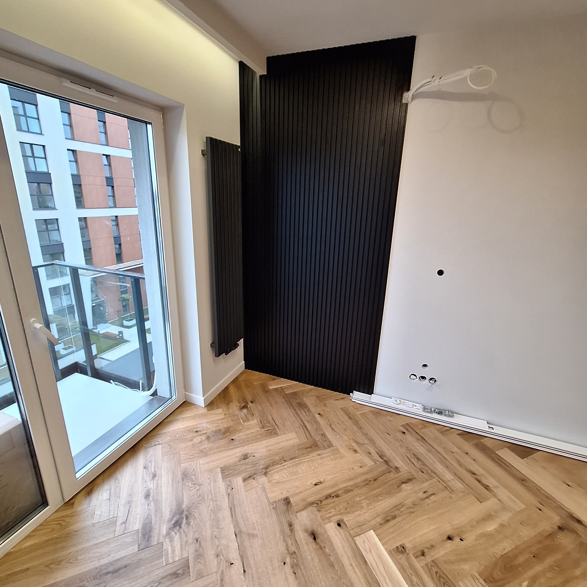 Nowoczesne wnętrze z drewnianą podłogą ułożoną w jodełkę, czarnymi lamelami na ścianie i grzejnikiem w tym samym kolorze, widok na balkon i budynek mieszkalny za oknem, widoczne przygotowane kable...