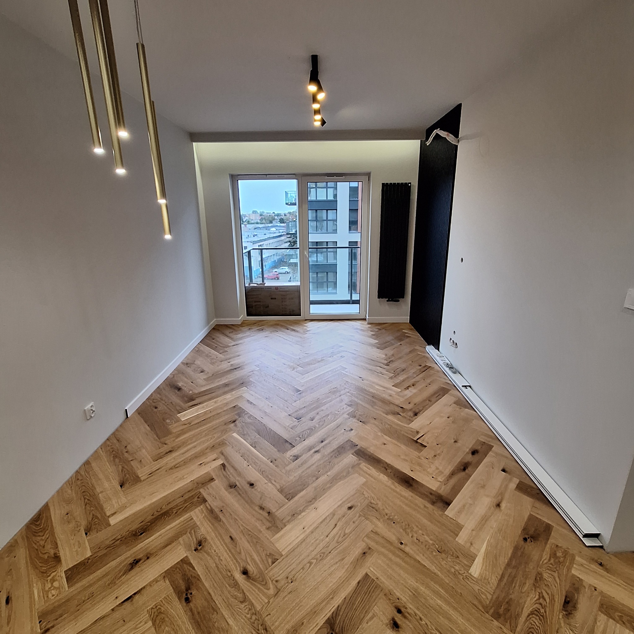 Parkiet ułożony w jodełkę w nowocześnie wykończonym pokoju z balkonem i designerskim oświetleniem sufitowym.