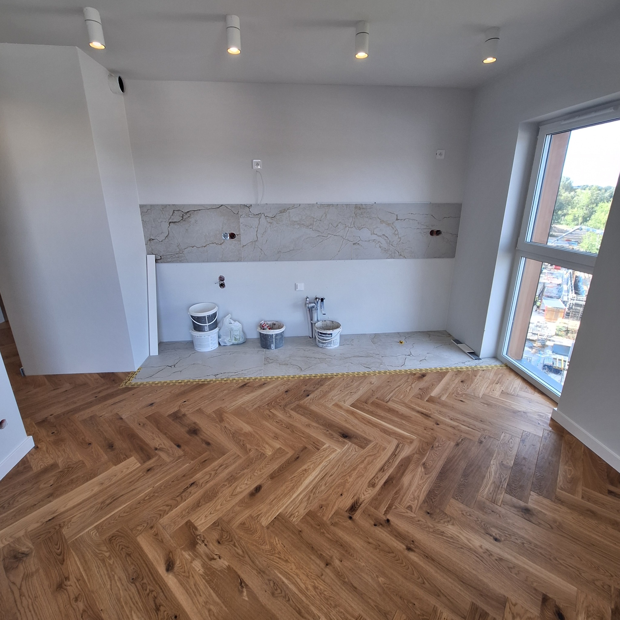 Przygotowana do montażu ściana z marmurowymi płytkami w nowym mieszkaniu, widoczne wiaderka z farbą i klejem, podłoga w jodełkę, okno z widokiem na budowę.