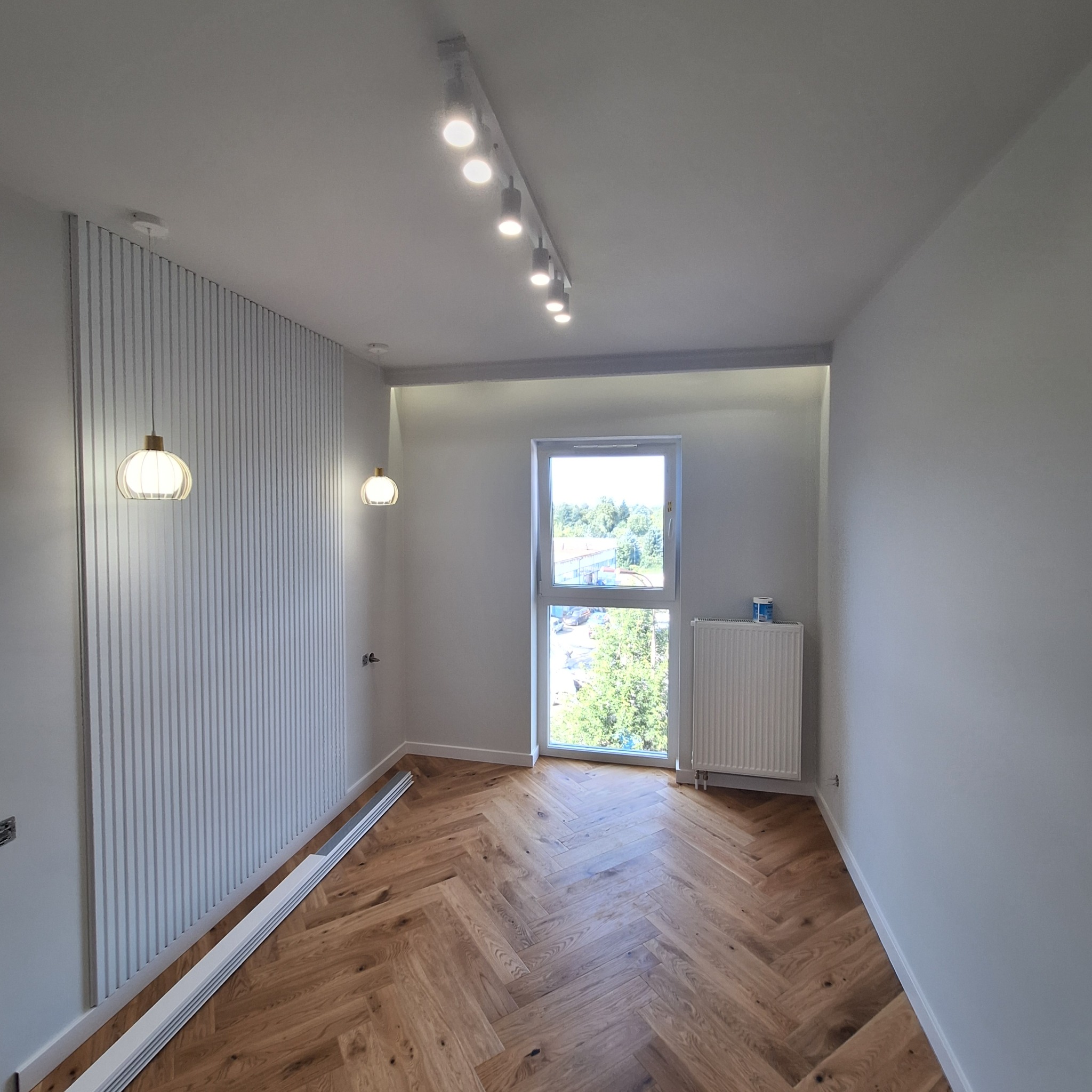 Jasne wnętrze z parkietem w jodełkę, dekoracyjną ścianą z lamelami, oświetleniem punktowym i dwoma wiszącymi lampami kulami.