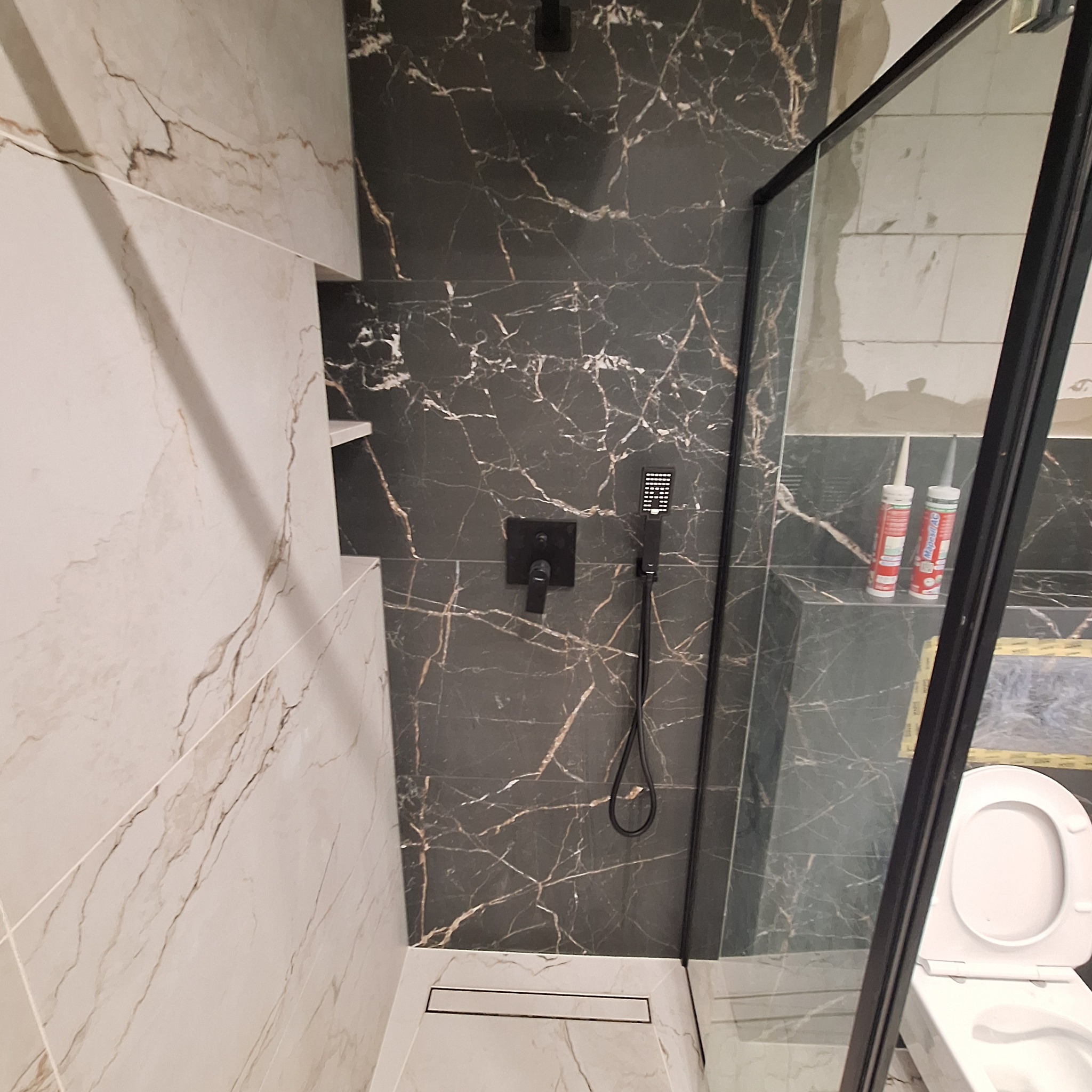Kabina prysznicowa z czarną armaturą i marmurowymi płytkami, widoczne tuby silikonu i toaleta za szybą